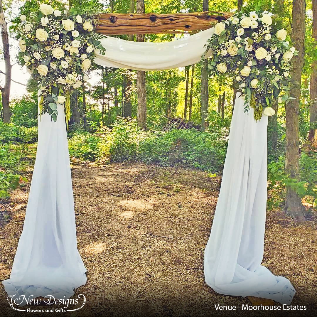 A large square arch in a wooded area, with white material and an all white flower installment in each corner.